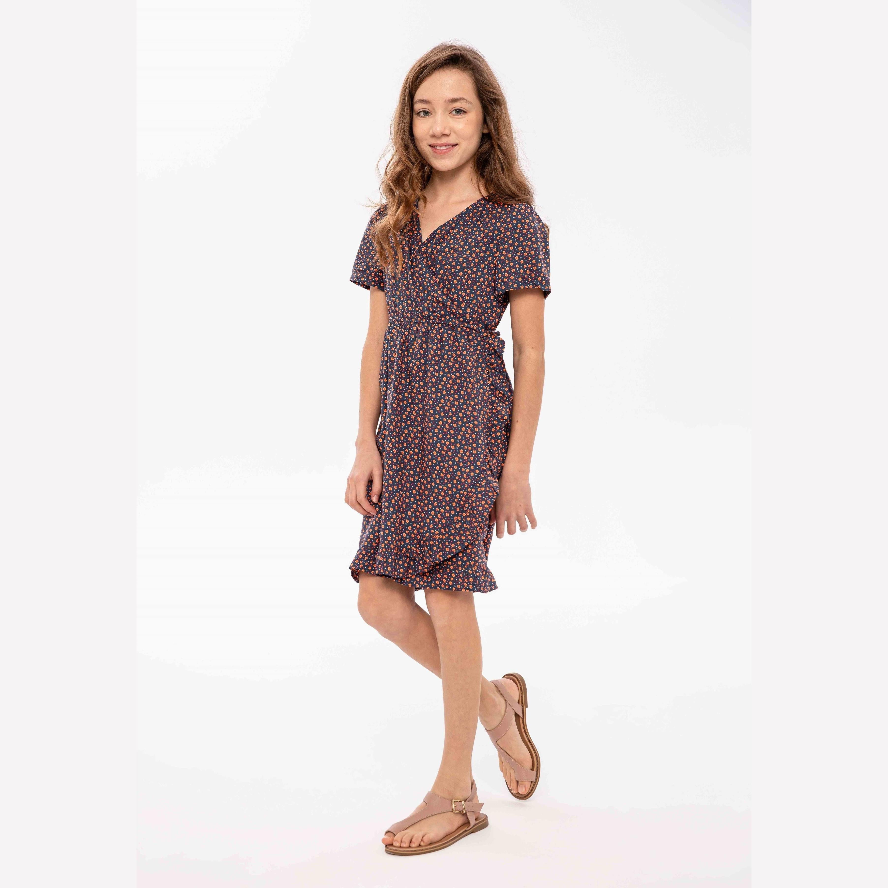 Young girl wearing navy floral wrap dress with short sleeves and ruffled hem, standing against white background wearing brown sandals