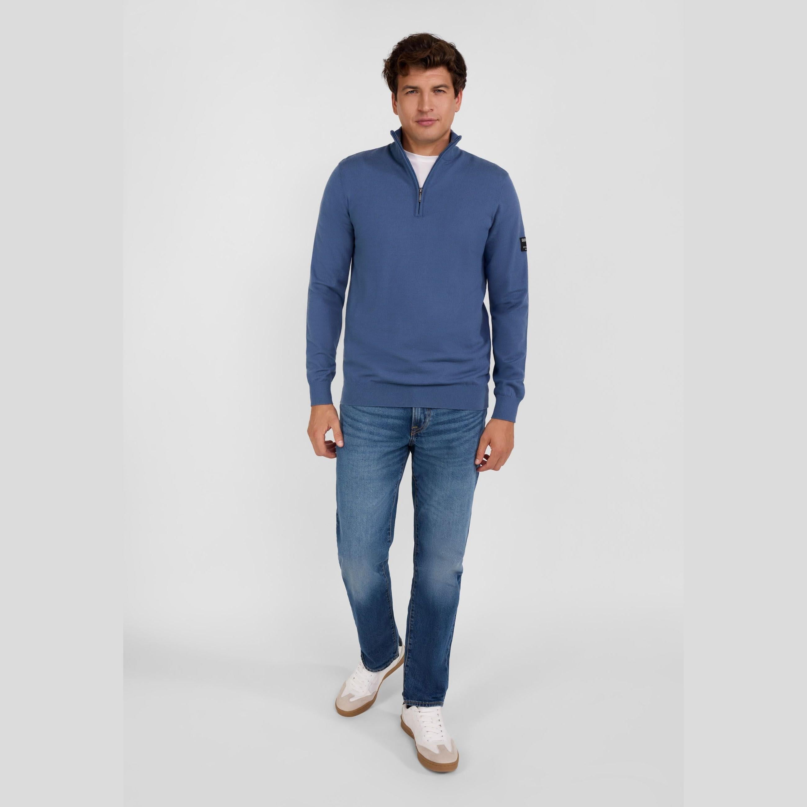 Men's blue half-zip stand collar jumper in a viscose blend, worn with jeans and white trainers.