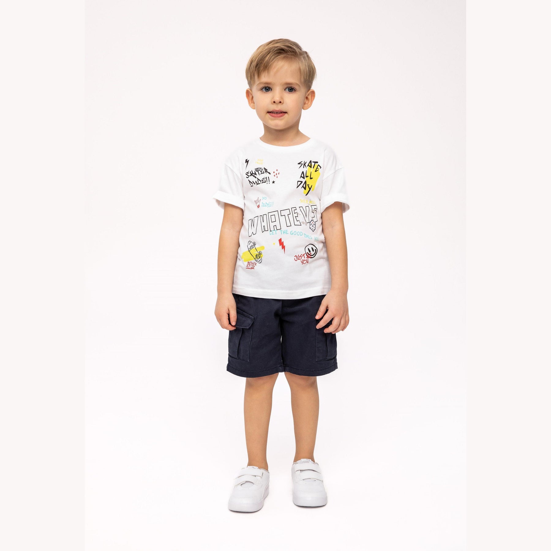 Young boy wearing white graphic t-shirt with skateboard print and navy shorts, standing against light background