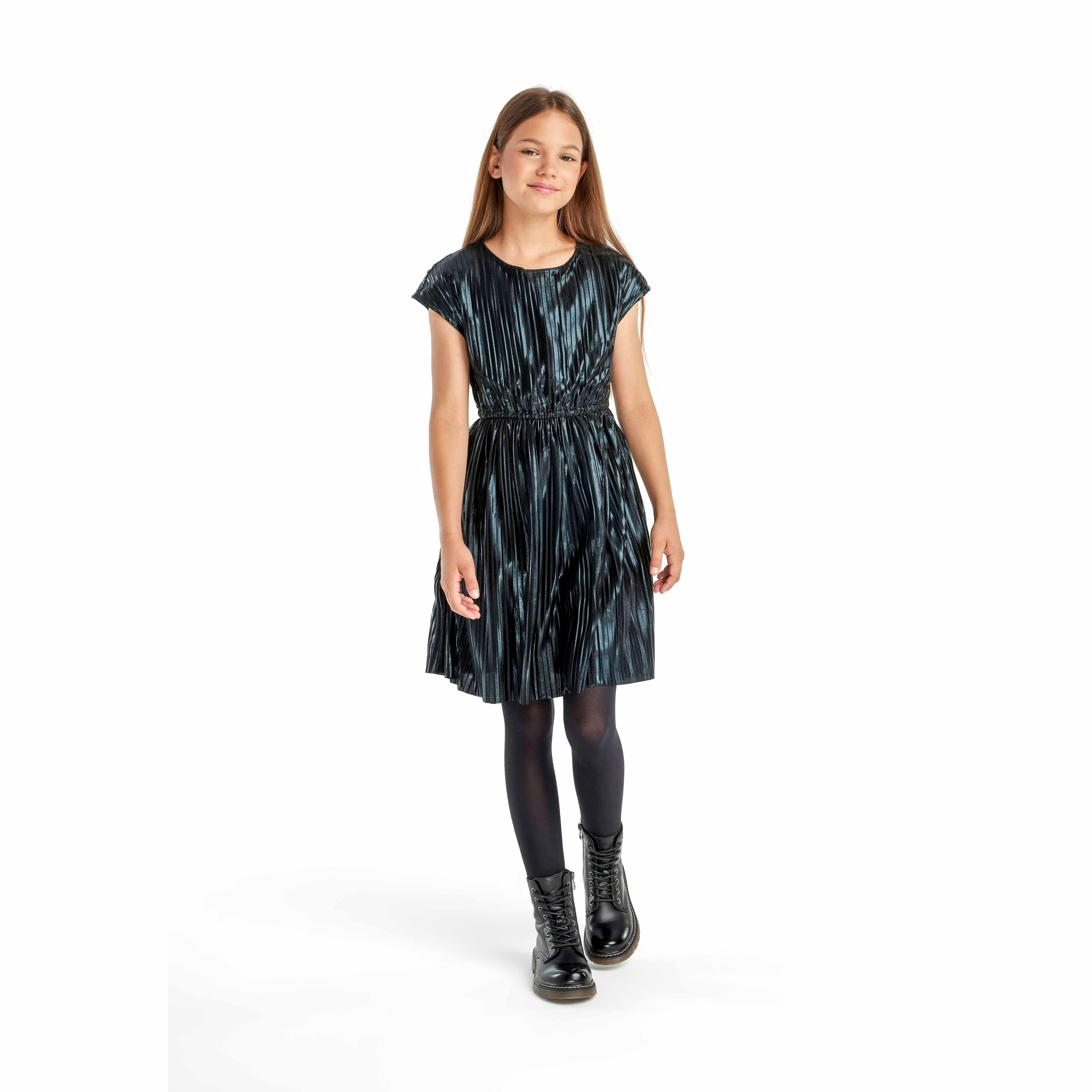 Young girl wearing metallic black pleated party dress with cap sleeves and black tights, smiling on white background